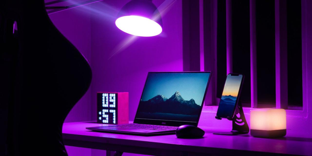 a laptop next to a mouse, phone, clock, and mouse, lit by purple lighting